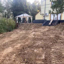 Przygotowany teren pod budowę z widocznymi śladami opon, częściowo ułożonymi schodami z bloczków betonowych i altaną w tle.