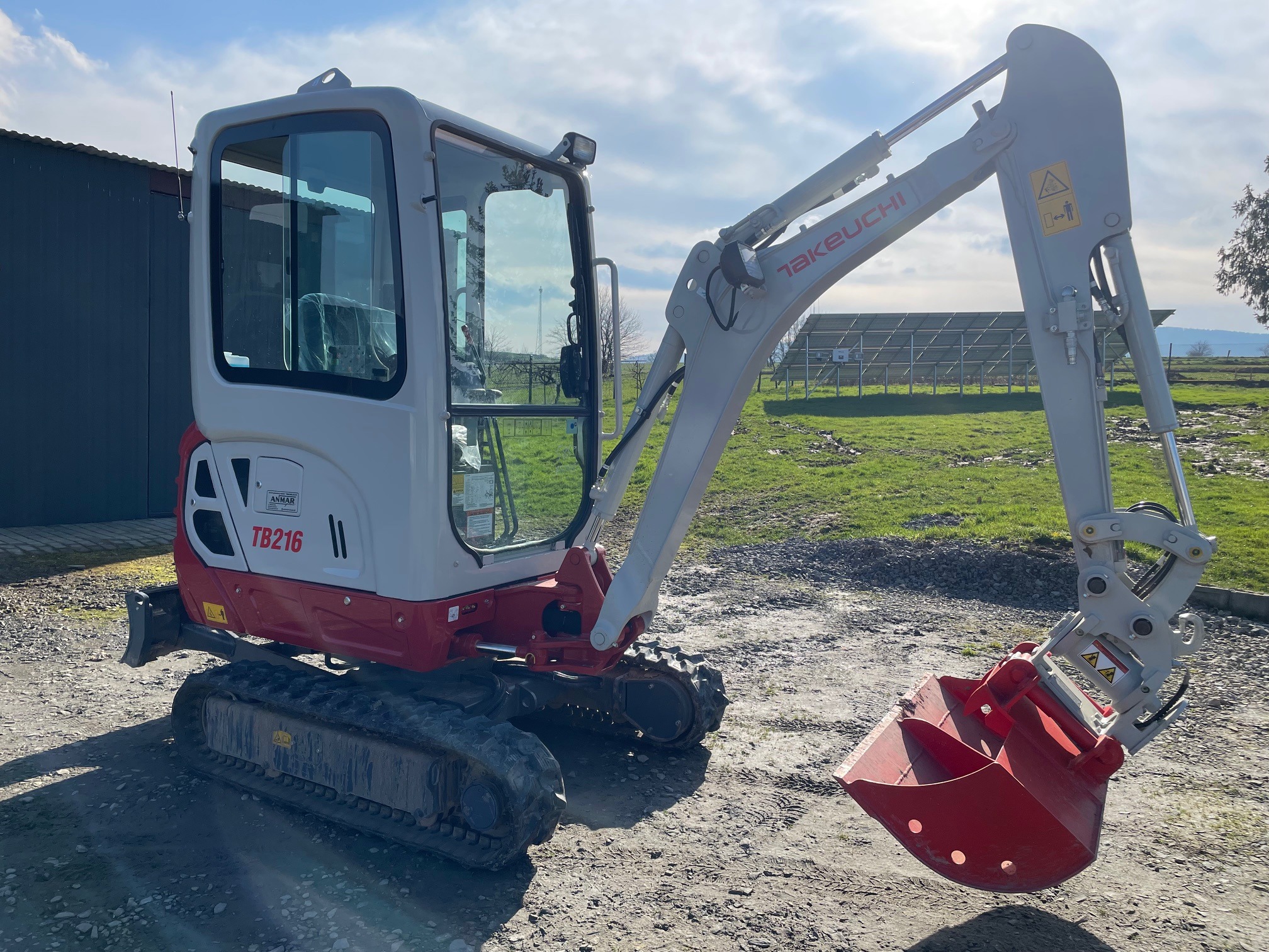 Kompaktowa, czerwono-biała minikoparka Takeuchi TB216 z łyżką, na tle zielonego terenu z panelami słonecznymi w oddali.