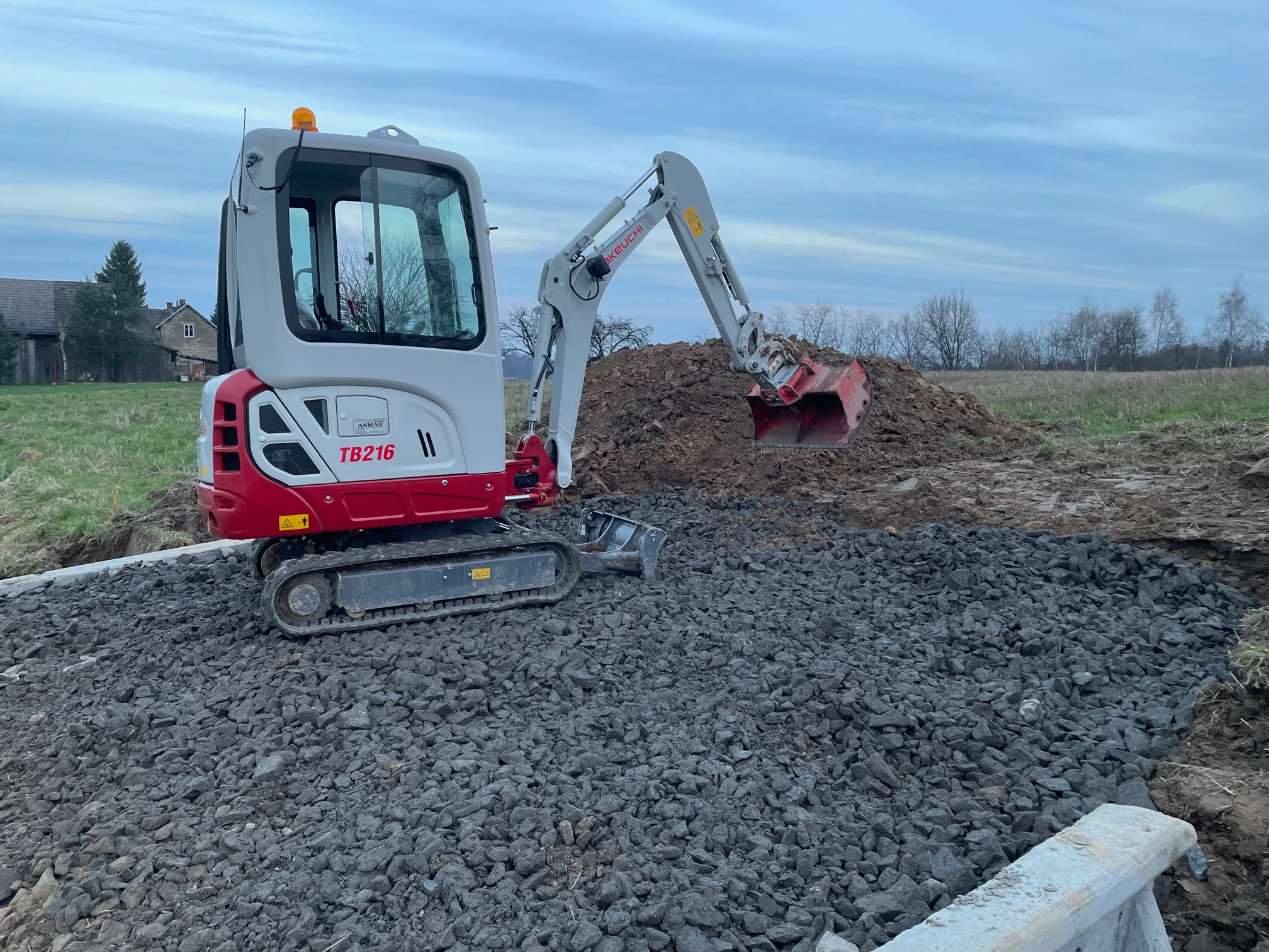 Mała koparka Takeuchi TB216 na żwirowym podłożu, z wykopaną ziemią w tle i widocznym fragmentem betonowego obrzeża.