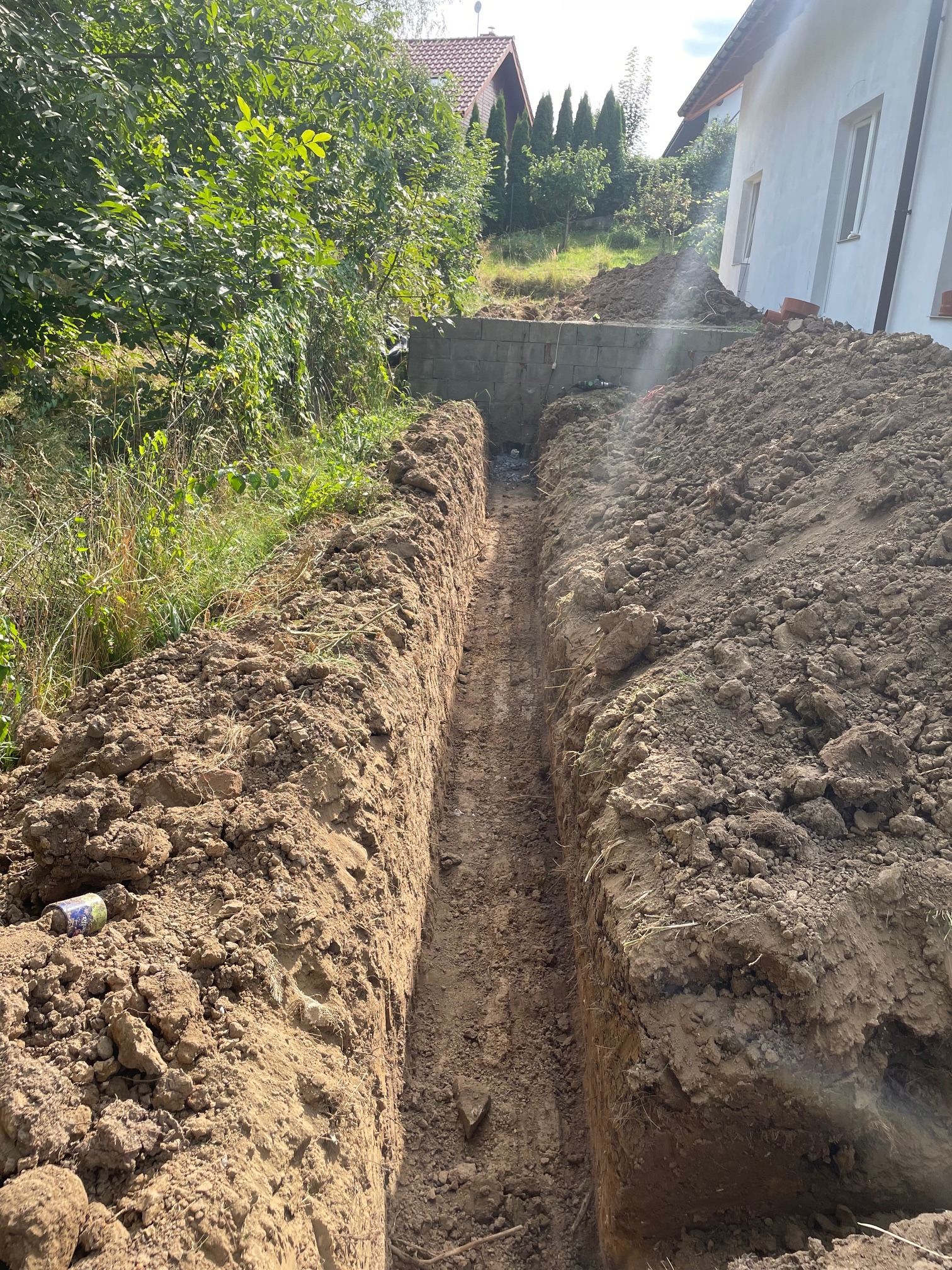 Wąski wykop w ziemi, widoczny z góry, z ziemią usypaną po bokach. W tle fragment muru oporowego z pustaków i budynek mieszkalny. Po lewej stronie leży puszka po napoju.