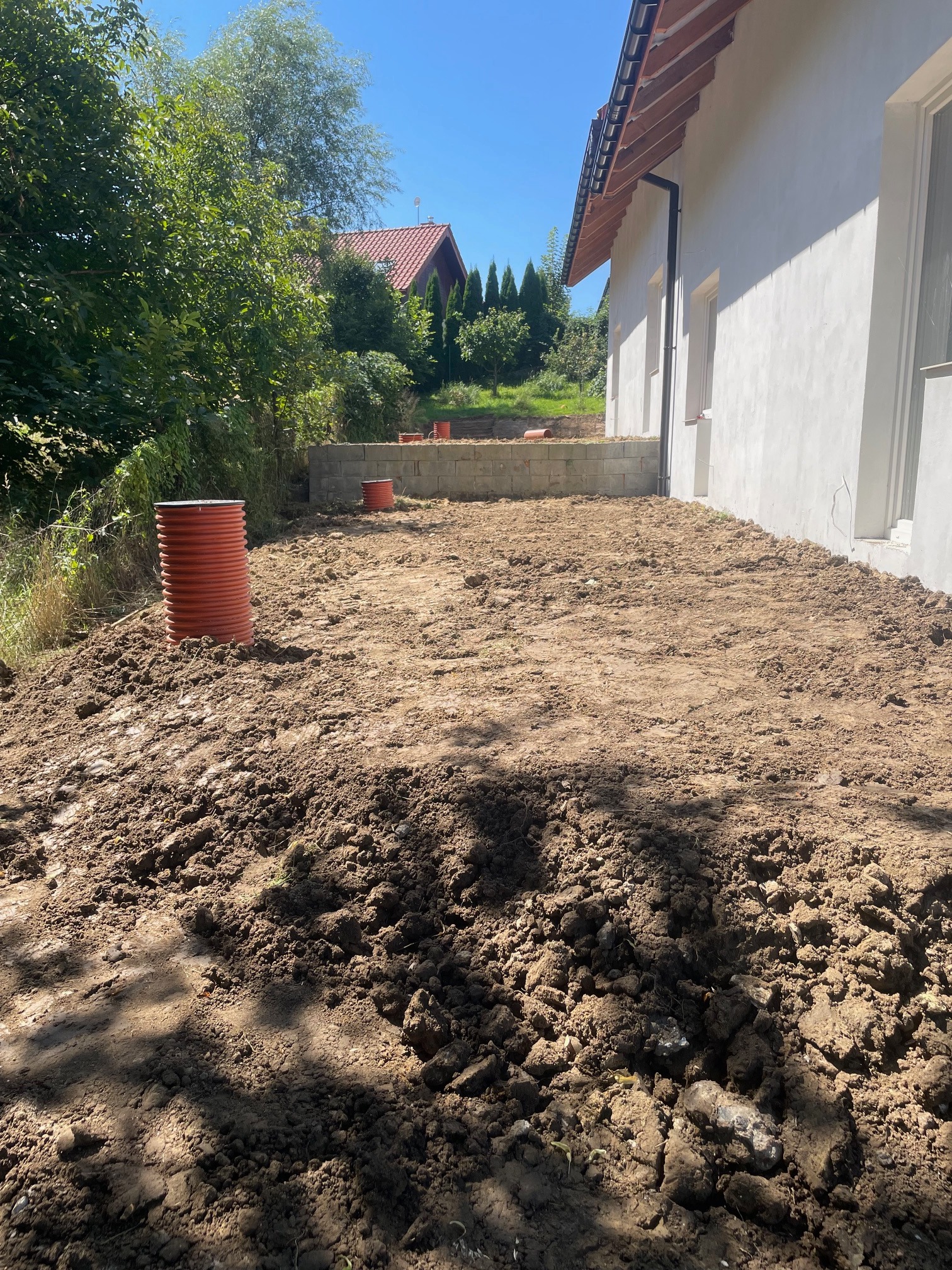 Przygotowany teren z ułożoną rurą drenarską w pobliżu budynku z białym tynkiem i widocznymi elementami konstrukcji ogrodzenia z betonowych bloczków.