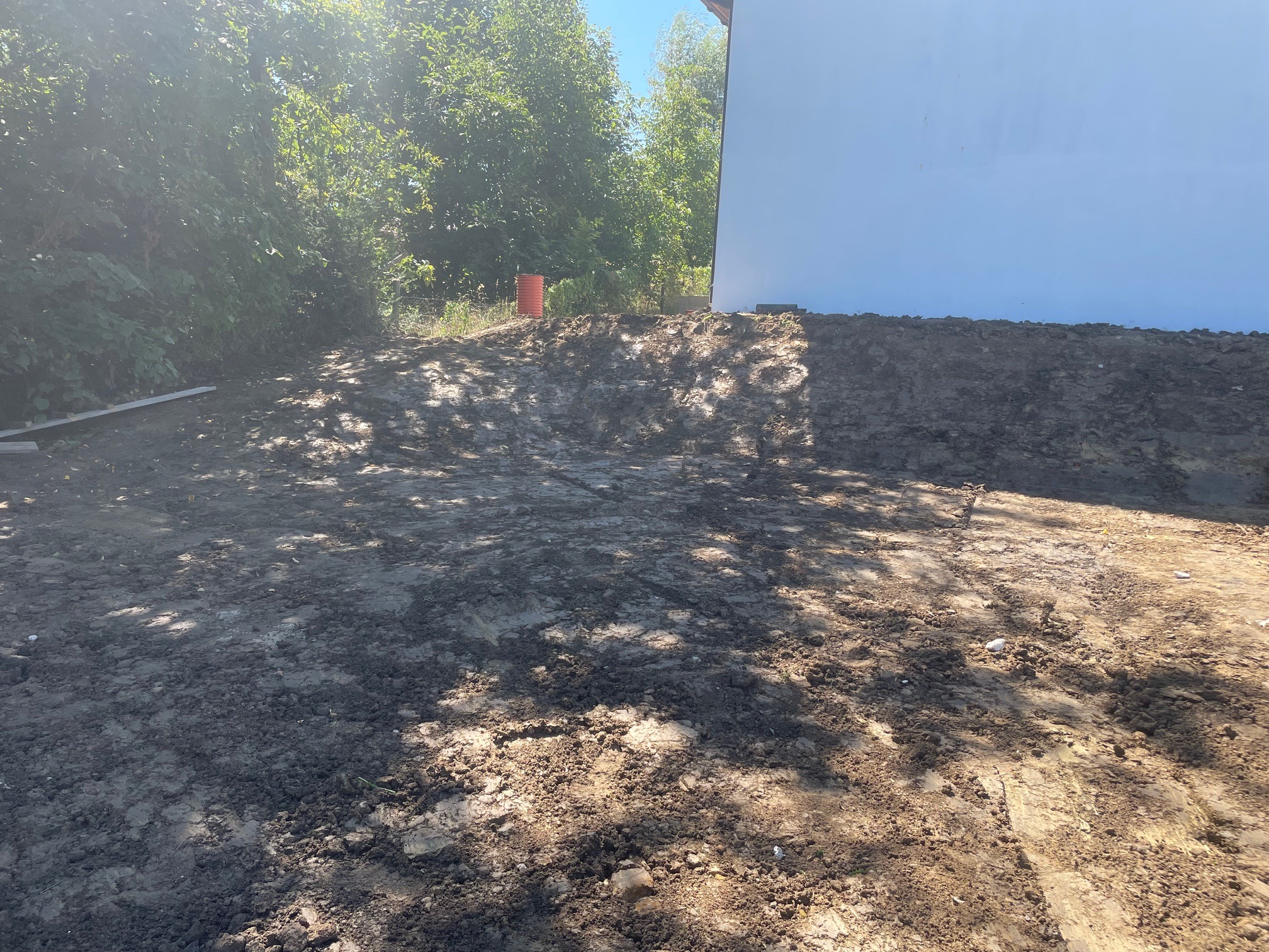 Przygotowany teren z pryzmą ziemi pod budowę, widoczny fragment budynku i drzewa w tle, pomarańczowy pojemnik na skraju pryzmy.