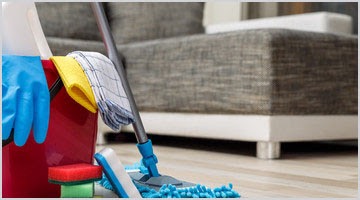 Mop z niebieskimi frędzlami oparty o ścianę, wiadro z płynami, szmatkami i gąbkami, niebieskie rękawice gumowe, fragment szarej sofy na białych nóżkach, podłoga z paneli.