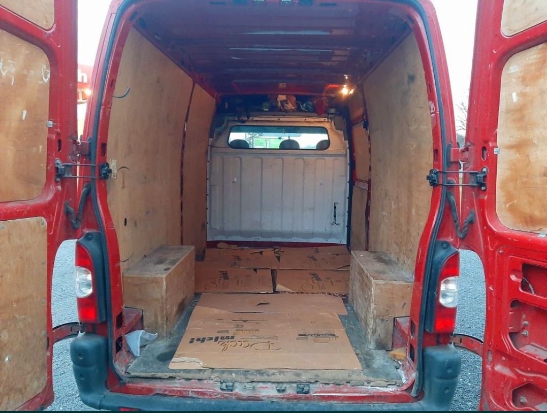 Wnętrze czerwonego furgonetki z otwartymi tylnymi drzwiami, wyłożone sklejką, z kartonami i drewnianymi skrzynkami na podłodze, widok z tyłu pojazdu.