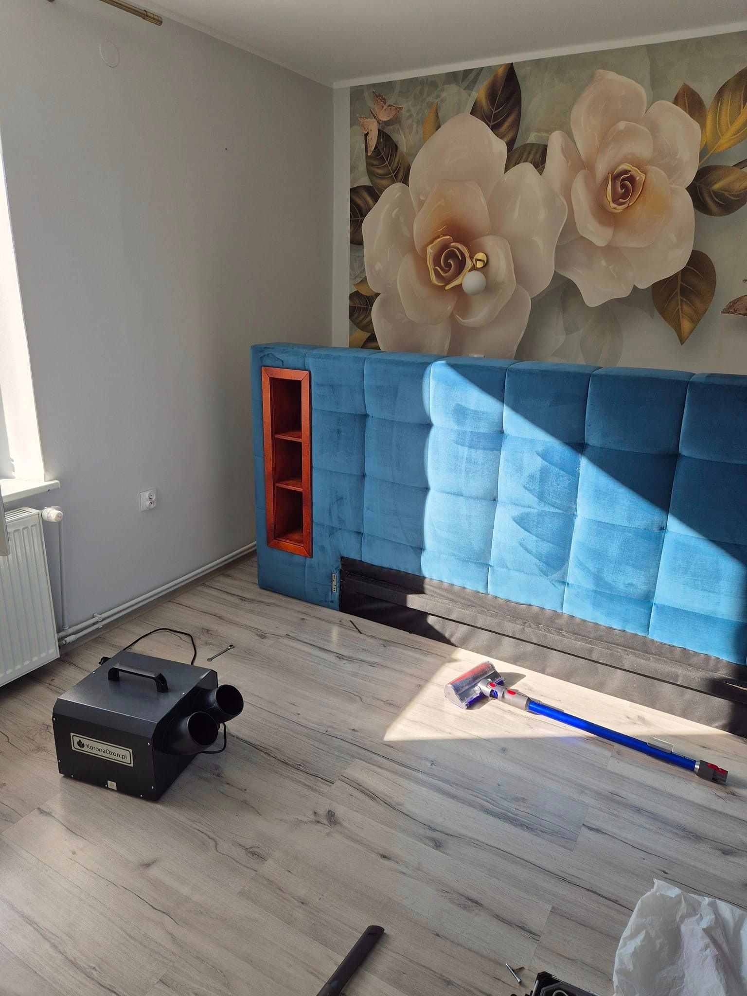 Ozonator KoronaOzon.pl, odkurzacz i niebieski tapicerowany zagłówek łóżka w pokoju z kwiatową tapetą, prawdopodobnie po zabiegu odkażania.