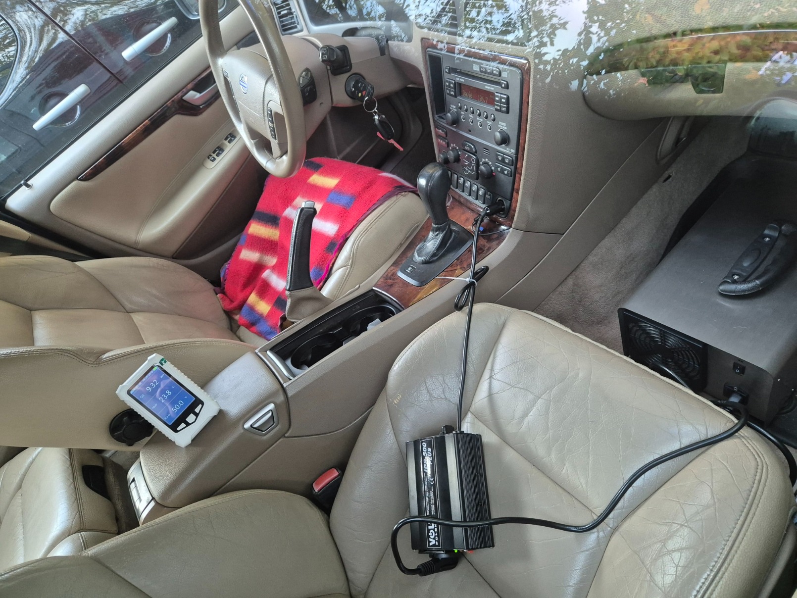 Wnętrze samochodu z widocznym ozonatorem podłączonym do zasilania, umieszczonym na fotelu pasażera, obok tester jakości powietrza na konsoli środkowej.