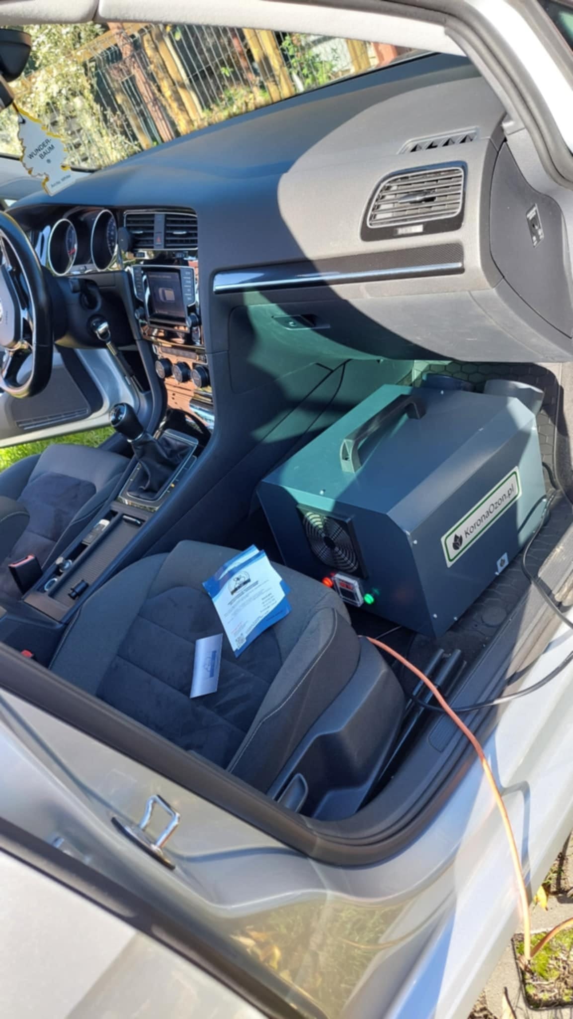 Wnętrze samochodu z ozonatorem KoronaOzon.pl na siedzeniu pasażera, widoczne ulotki informacyjne oraz kabel zasilający. Urządzenie stoi na ciemnoszarym fotelu obok otwartych drzwi.