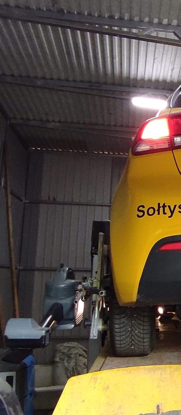 Żółty samochód osobowy na podnośniku w warsztacie, podczas ustawiania geometrii kół. Widoczne urządzenie do pomiaru i regulacji z przodu i z tyłu koła.