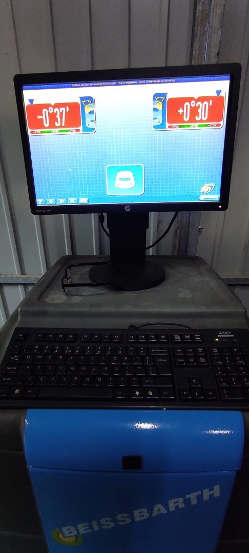 Monitor z oprogramowaniem do ustawiania geometrii kół pojazdu, klawiatura i okulary leżące na stanowisku diagnostycznym.