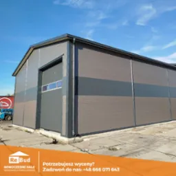 Zewnętrzny widok hali magazynowej z płyt warstwowych w odcieniach szarości i brązu, z częściowo otwartą bramą segmentową, na betonowym placu pod błękitnym niebem. W tle widoczny fragment czerwonej...