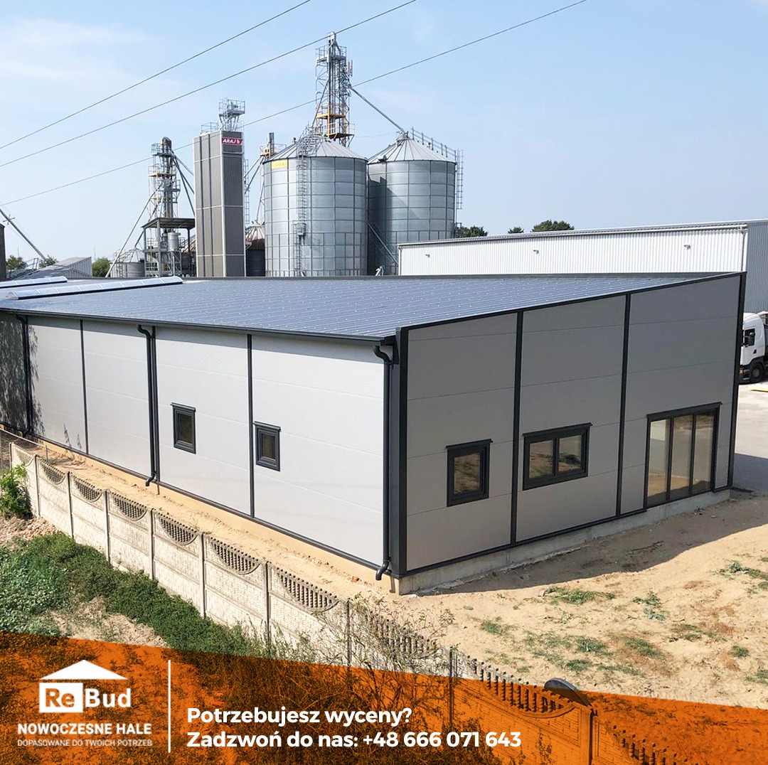 Nowoczesna hala magazynowa z szarych płyt warstwowych z ciemną stolarką okienną i dachem, na tle silosów zbożowych i błękitnego nieba.