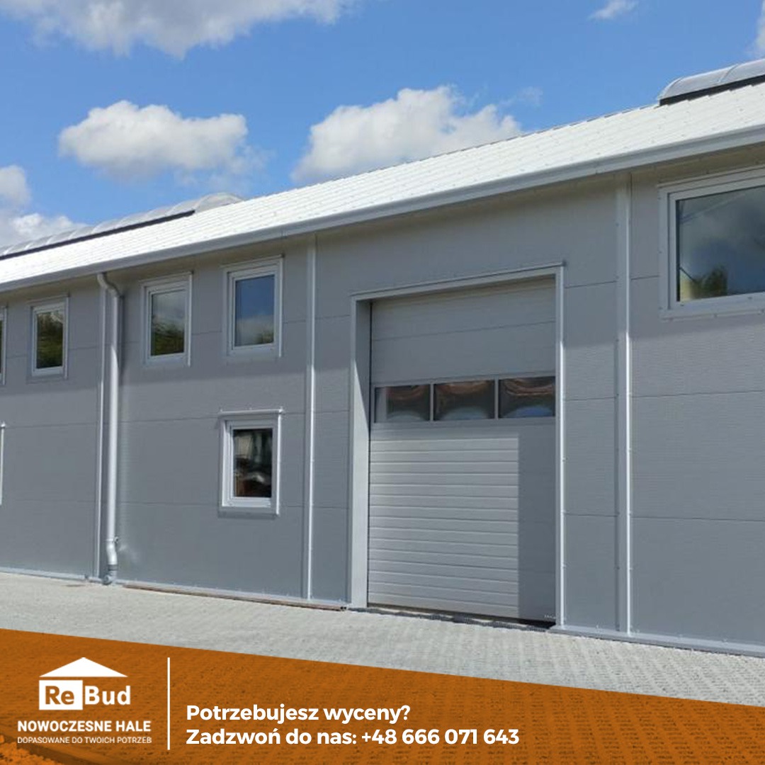 Gotowa hala przemysłowa z szarych płyt warstwowych z częściowo otwartą bramą wjazdową, widoczne okna i rynny, błękitne niebo w tle.