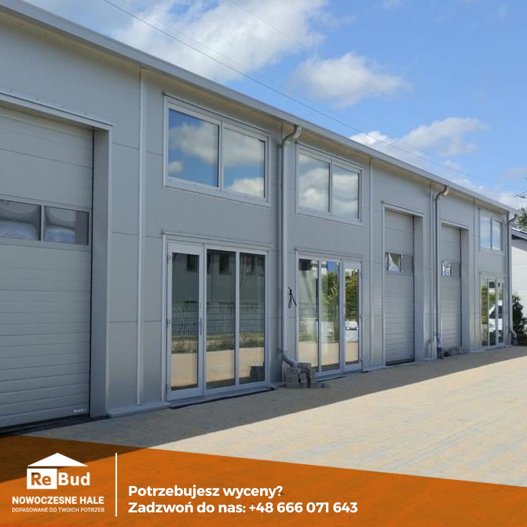 Nowoczesna hala przemysłowa z szarych płyt warstwowych z dużymi oknami i bramami segmentowymi w słoneczny dzień, widoczny fragment reklamy firmy.