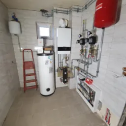 Wnętrze kotłowni z nową instalacją: biały piec gazowy, czerwony zbiornik wyrównawczy, biały zasobnik ciepłej wody owinięty folią, rozdzielacz podłogowy w zabudowie, liczne rury i zawory, drabina...