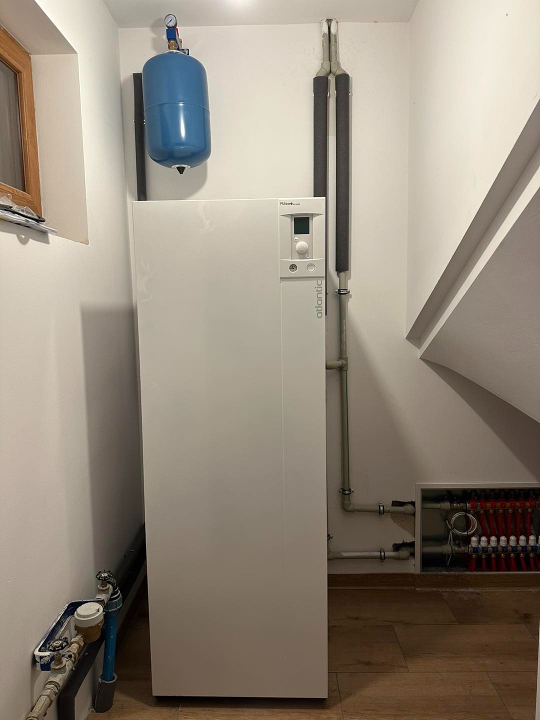 Biały piec grzewczy Atlantic z niebieskim zbiornikiem wyrównawczym na ścianie, widoczne rury CO z izolacją oraz rozdzielacz podłogowy w ścianie.