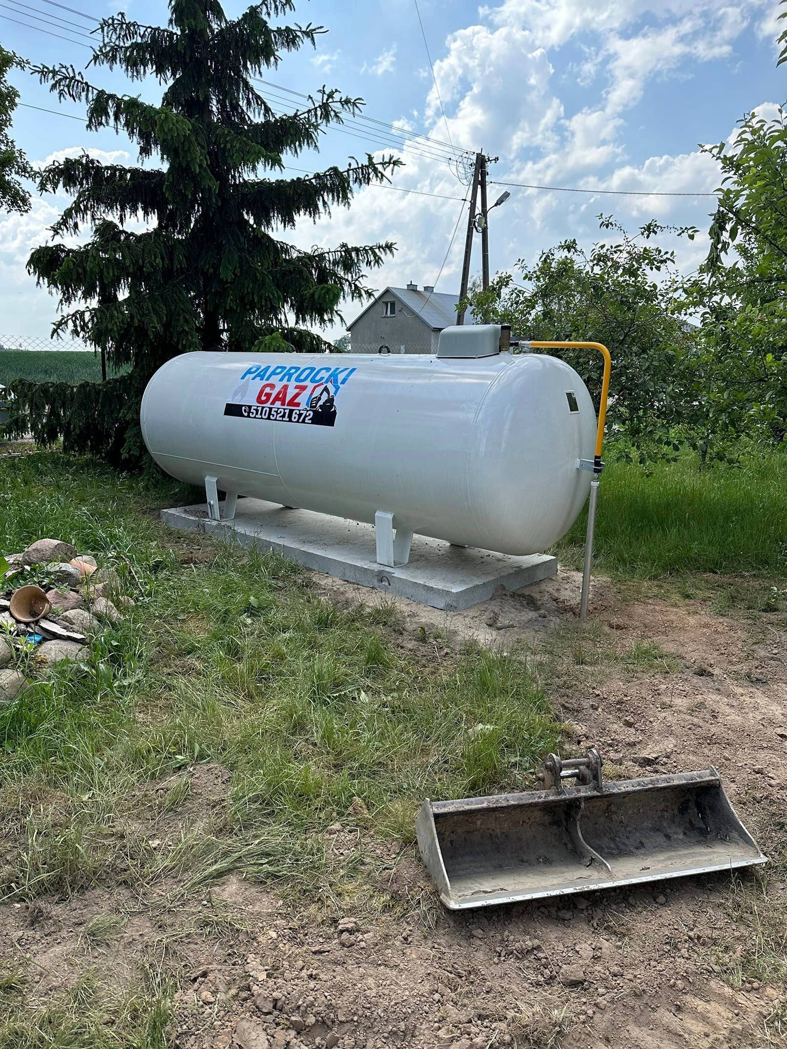 Biały zbiornik na gaz LPG z żółtym przyłączem, umieszczony na betonowej podstawie w trawiastym terenie z widocznym łyżką koparki i budynkiem w tle.