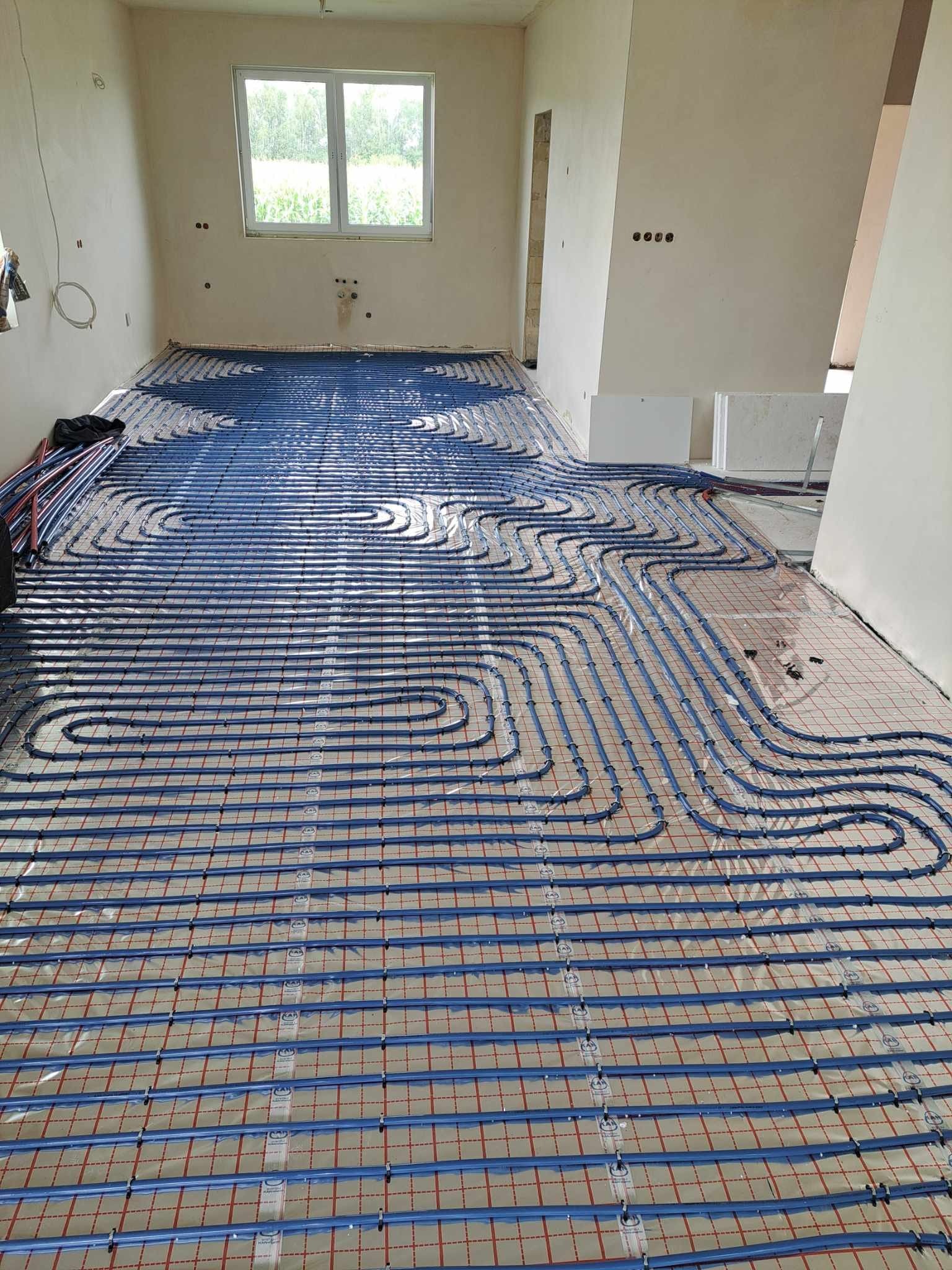 Niebieskie rury ogrzewania podłogowego ułożone na siatce w surowym pomieszczeniu z oknem i instalacją elektryczną na ścianach.