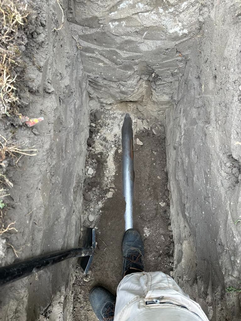 Wykop z rurą, widok z góry, obok łopata i buty osoby wykonującej pracę.