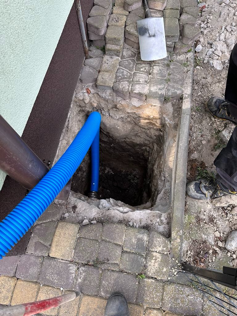 Wykop pod fundamentem budynku z widoczną niebieską rurą karbowaną, prawdopodobnie osłonową, prowadzącą w głąb wykopu. Obok wykopu leżą kostki brukowe i łopata.