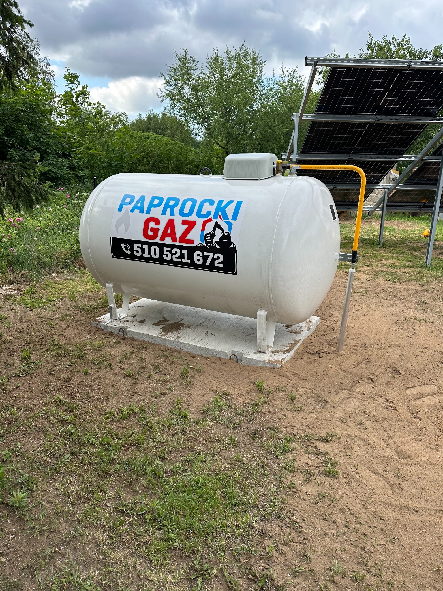 Biały zbiornik gazowy z niebiesko-czerwonym napisem 'PAPROCKI GAZ' i numerem telefonu, umieszczony na betonowej podstawie na trawiastym terenie, z panelami słonecznymi w tle.