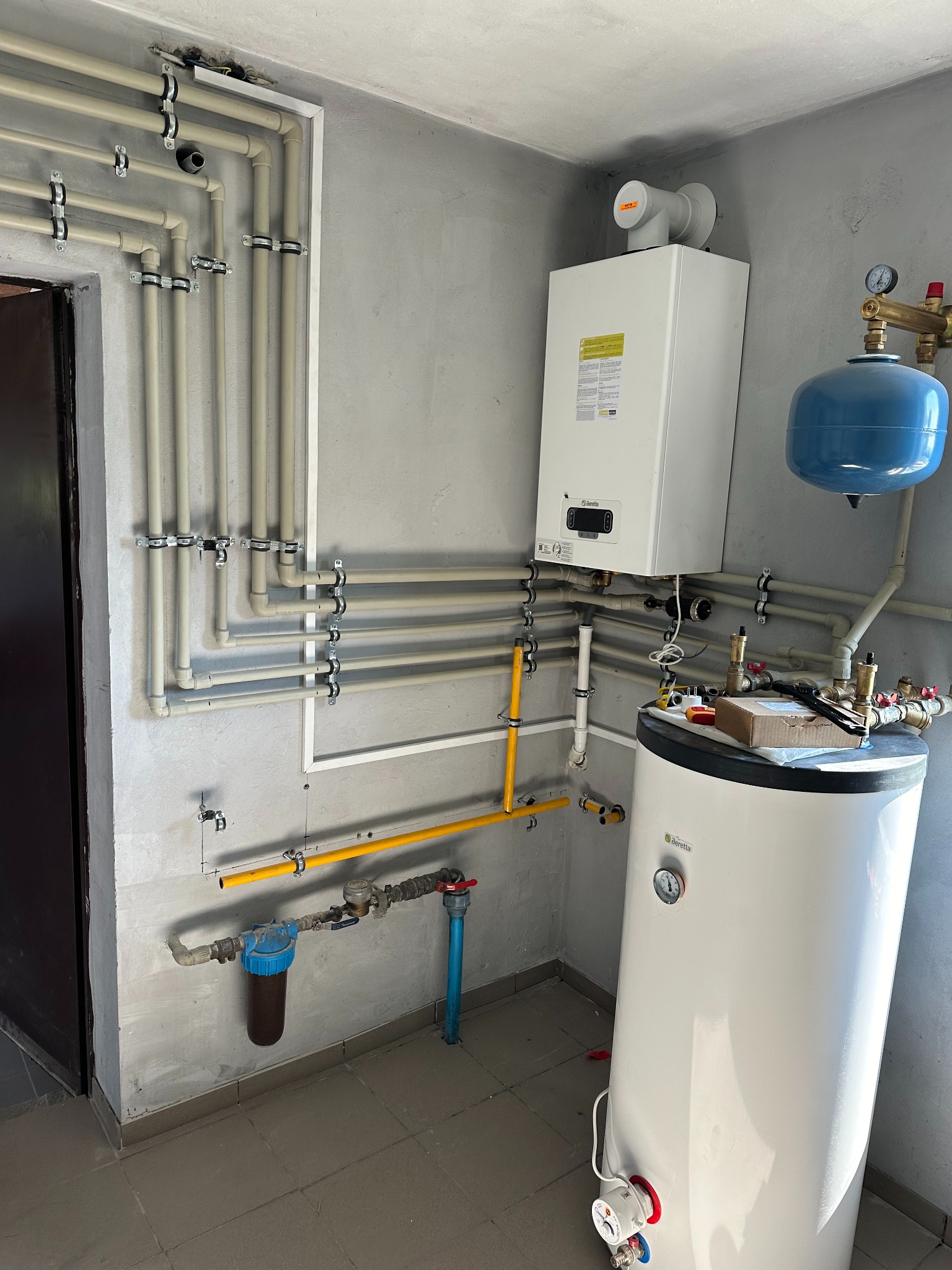 Nowoczesna kotłownia: biały piec gazowy Beretta, zbiornik na wodę, rozbudowana instalacja rurowa z licznymi mocowaniami na surowej ścianie, widoczne narzędzia na zbiorniku.