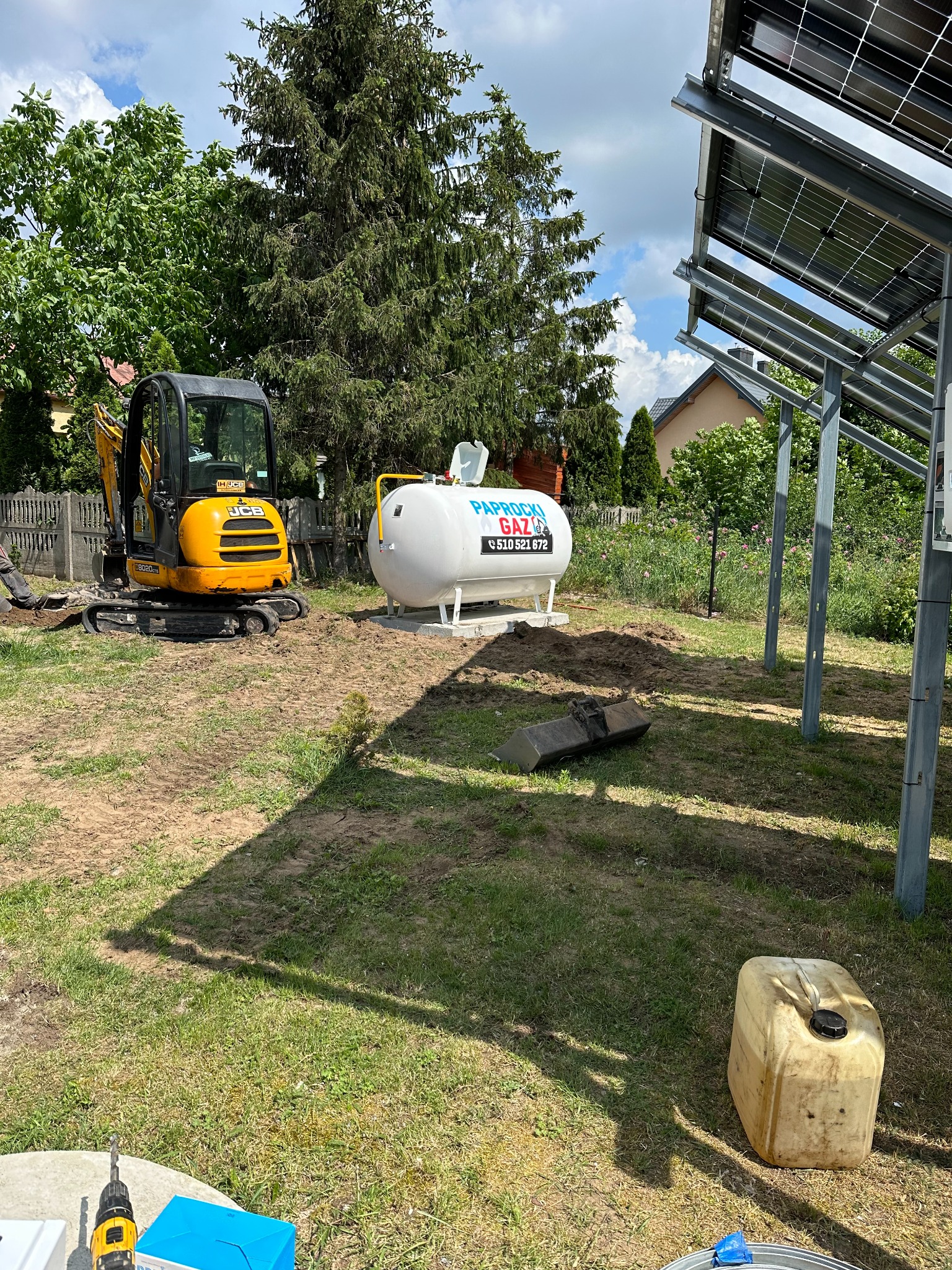 Biały zbiornik na gaz z naklejką informacyjną, żółta mini koparka JCB i konstrukcja paneli fotowoltaicznych na trawiastym terenie w słoneczny dzień.