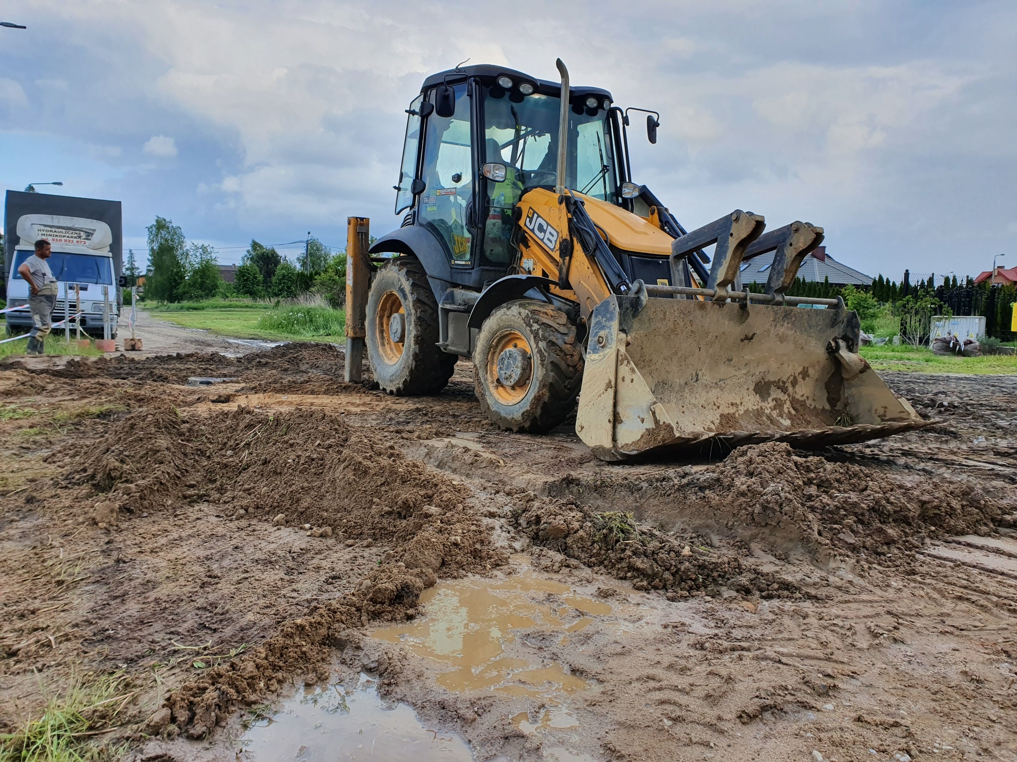 Żółto-czarna koparko-ładowarka JCB na placu budowy z błotnistym terenem, widoczna ciężarówka w tle i pracownik