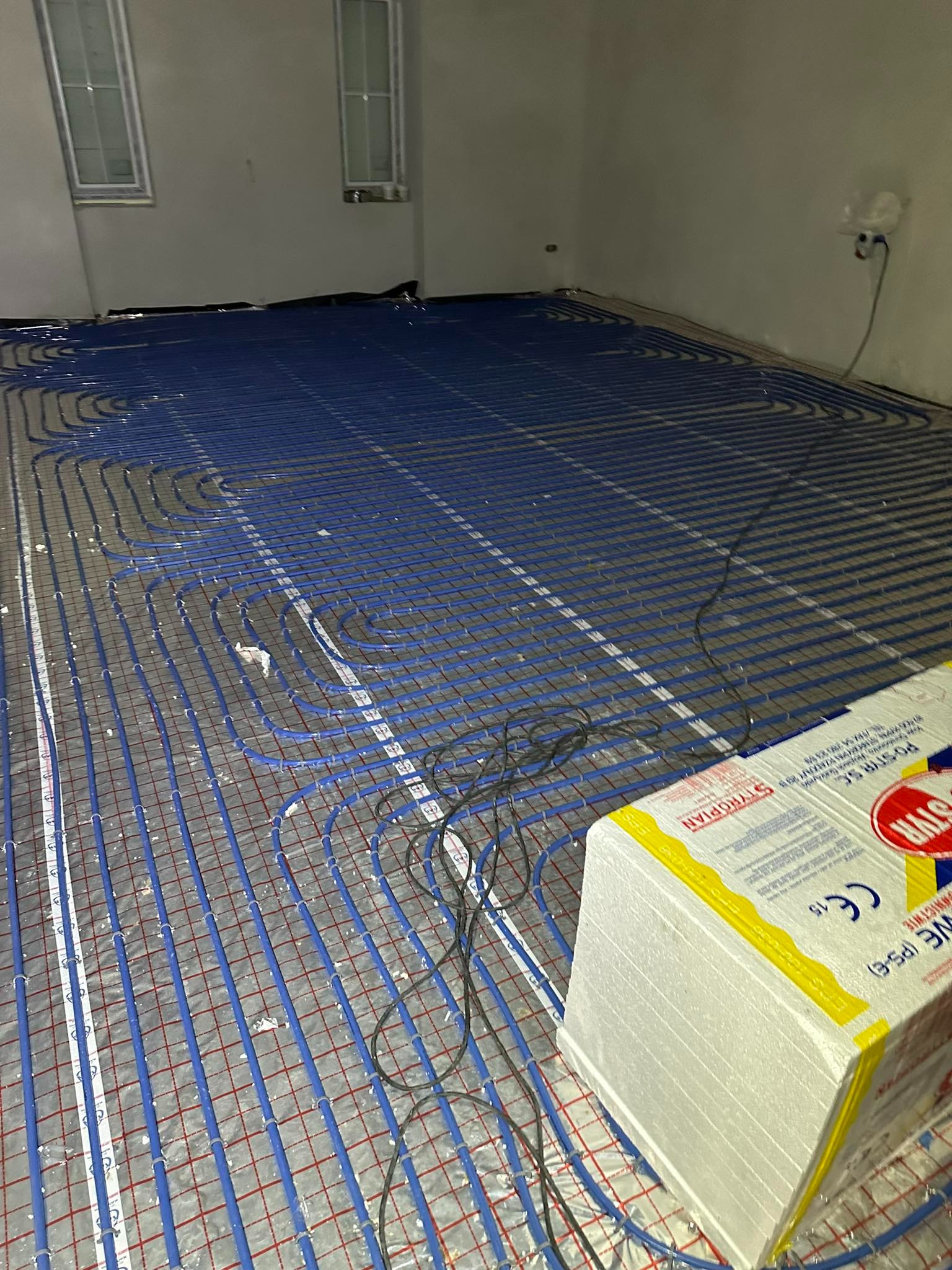 Niebieskie rury ogrzewania podłogowego rozłożone na siatce, widoczne okna w tle i styropianowe płyty izolacyjne w rogu pomieszczenia, etap instalacji.