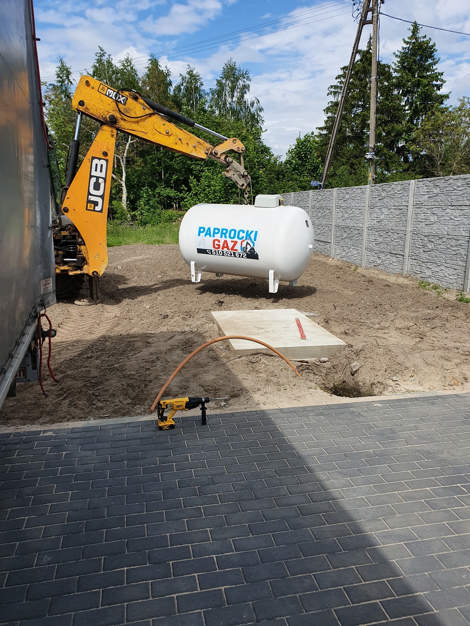 Montaż zbiornika gazowego na posesji: biały zbiornik z logo firmy, żółta koparka JCB, wiertarka udarowa DeWalt na kostce brukowej, wykopany dół w ziemi.