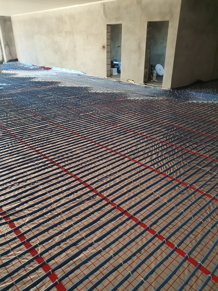 Rozłożona instalacja ogrzewania podłogowego z rurkami na folii z czerwoną siatką w surowym stanie, widoczne przygotowane otwory drzwiowe w tle.