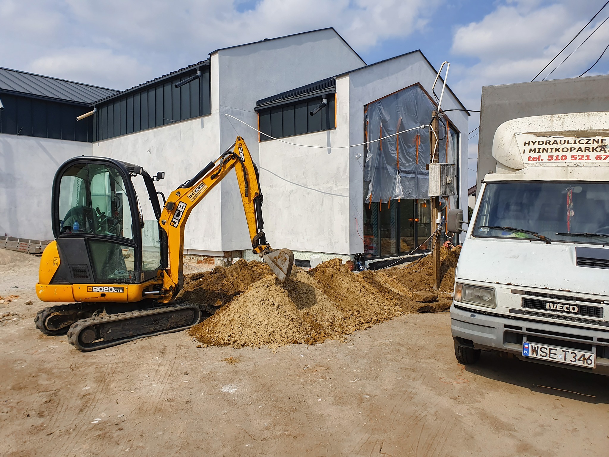 Żółta mini koparka JCB podczas prac ziemnych przy budynku w trakcie budowy, obok zaparkowany biały samochód dostawczy z napisem 'hydrauliczne i minikoparka'.