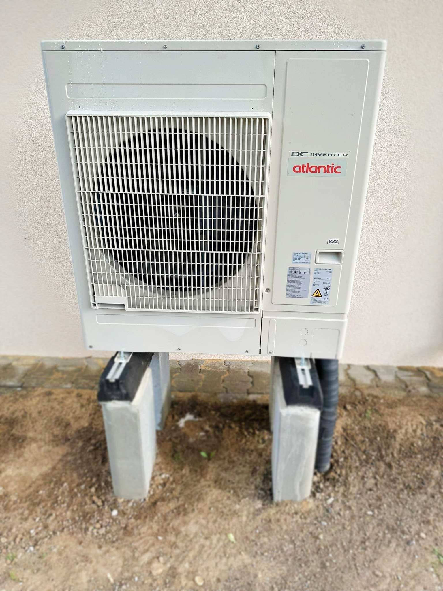 Zewnętrzna jednostka pompy ciepła Atlantic DC Inverter zamontowana na betonowych podstawach z gumowymi podkładkami, widoczna tabliczka znamionowa i oznaczenie R32.