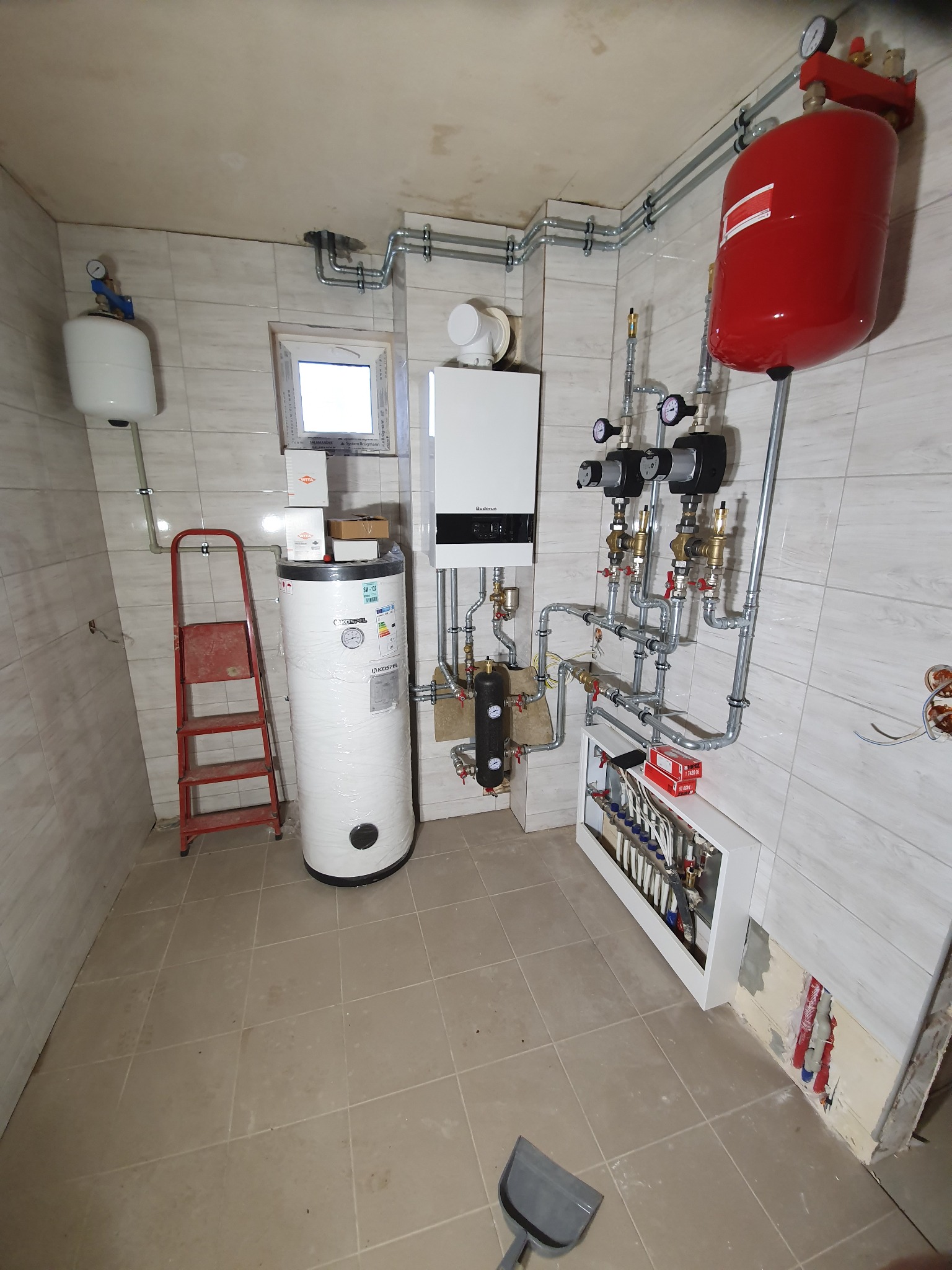 Wnętrze kotłowni z nową instalacją: biały piec gazowy, czerwony zbiornik wyrównawczy, biały zasobnik ciepłej wody owinięty folią, rozdzielacz podłogowy w zabudowie, liczne rury i zawory, drabina...