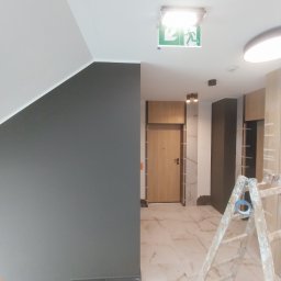 Krepis - Klatka schodowa w trakcie malowania, ściana w odcieniach szarości, marmurowa podłoga i aluminiowa drabina z niebieską taśmą malarską.