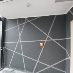 Krepis - Szara ściana biura z geometrycznym wzorem z białych linii, pomarańczowa skrzynka alarmowa, widoczny fragment sufitu z oświetleniem.