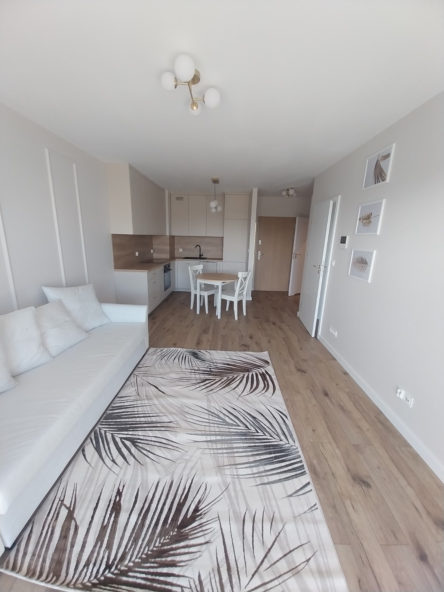 Jasne wnętrze mieszkania z aneksem kuchennym, białym narożnikiem i dywanem w liście palmy, drewniana podłoga, minimalistyczne obrazy na ścianie.