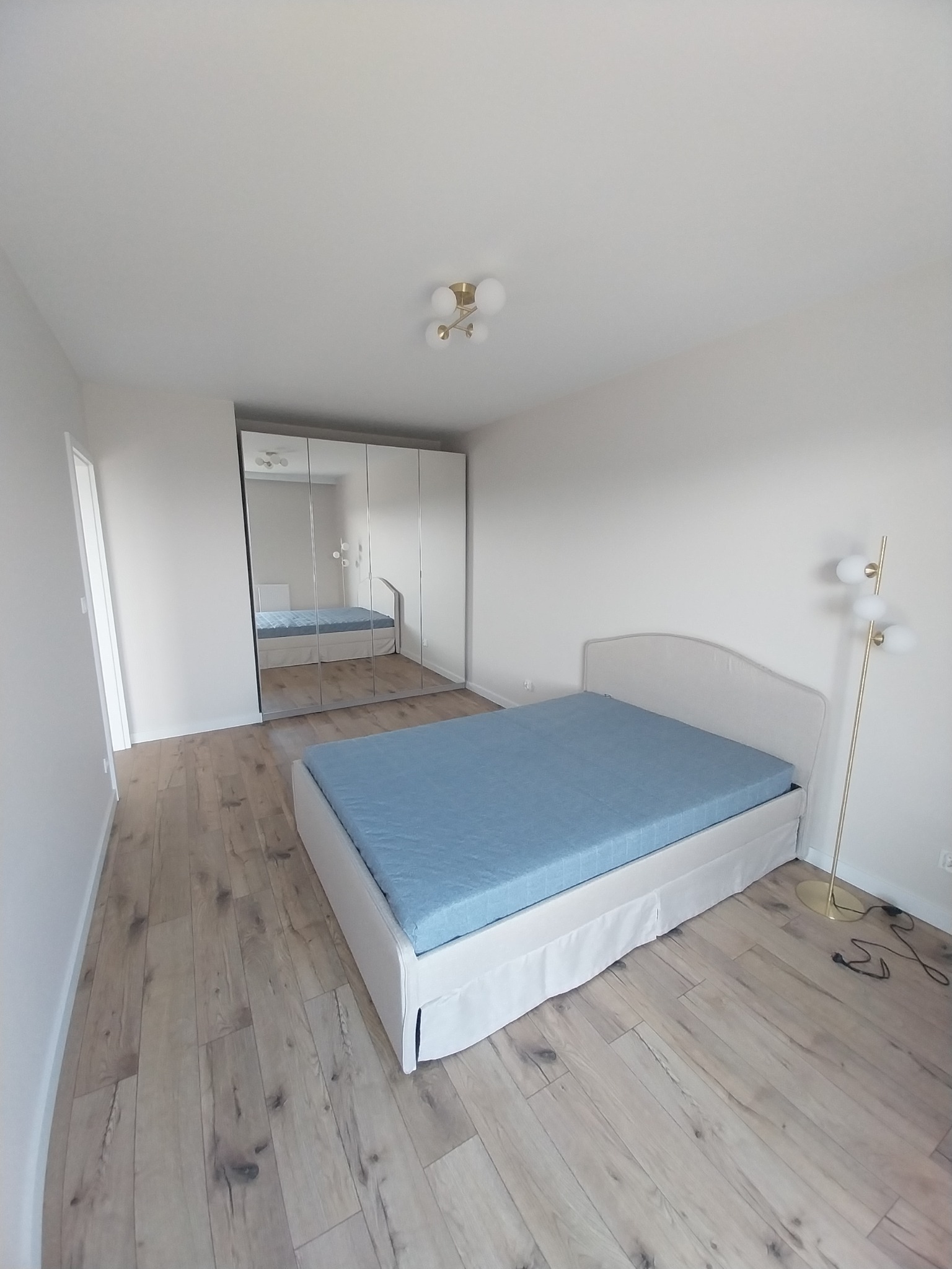 Jasna sypialnia z drewnianą podłogą, łóżkiem z błękitną pościelą i szafą z lustrzanymi drzwiami odbijającą wnętrze pokoju, oświetlona lampą podłogową z kulistymi kloszami.