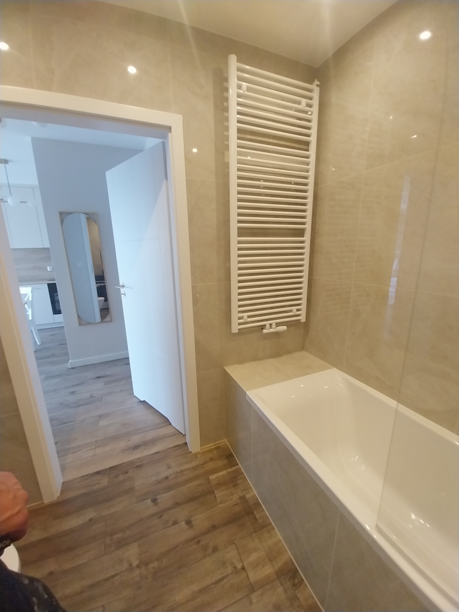 Jasna łazienka po liftingu z beżowymi płytkami, białą wanną z parawanem i grzejnikiem drabinkowym; widok na kuchnię przez otwarte drzwi.