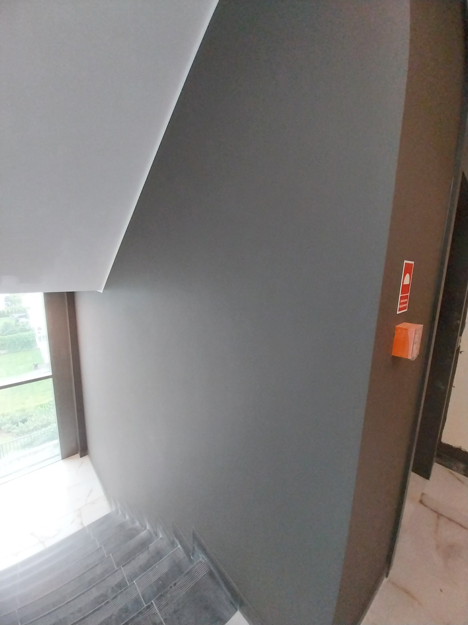 Świeżo pomalowana szara ściana klatki schodowej z widocznym przyciskiem alarmowym, marmurowe schody i okno z widokiem na zieleń.