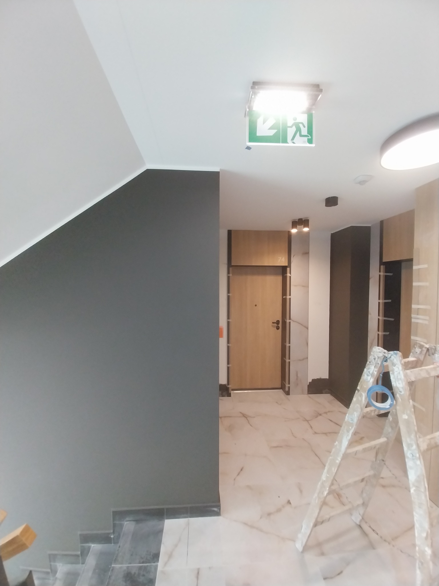 Klatka schodowa w trakcie malowania, ściana w odcieniach szarości, marmurowa podłoga i aluminiowa drabina z niebieską taśmą malarską.