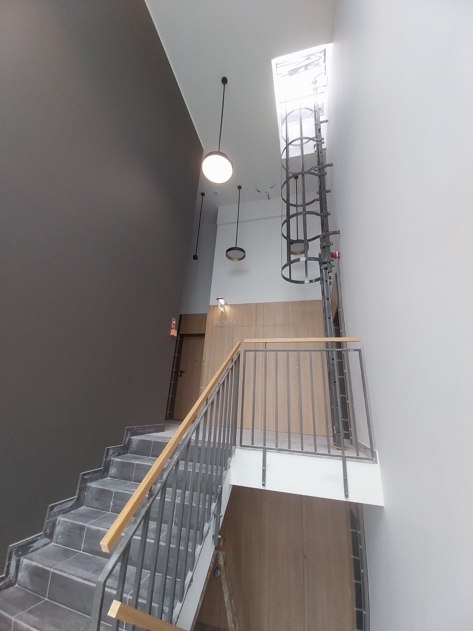 Wnętrze klatki schodowej z szarymi schodami wyłożonymi płytkami, drewnianą balustradą i przeszklonym szybem windowym w górnej części, oświetlone wiszącymi lampami i światłem dziennym