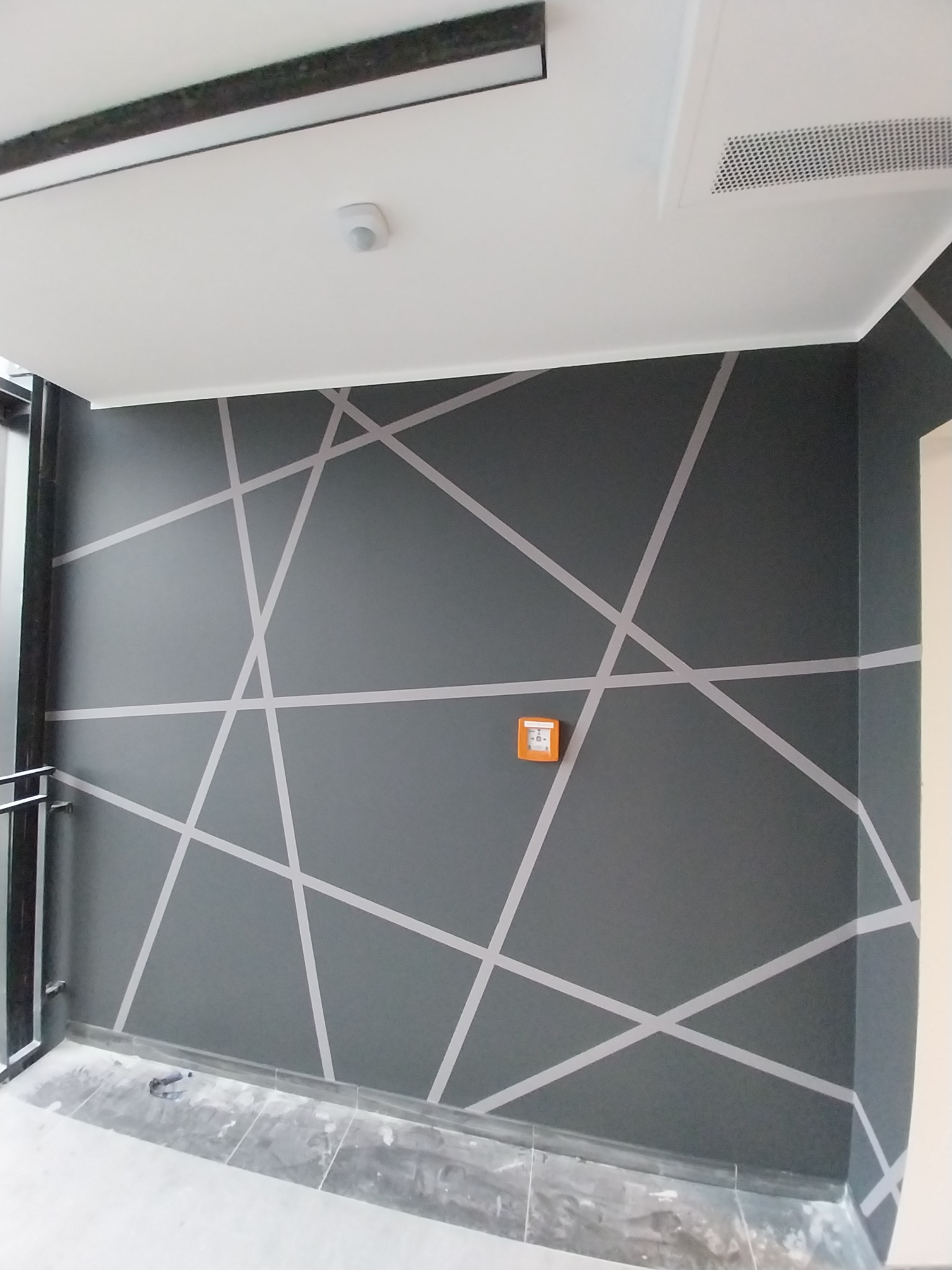 Szara ściana biura z geometrycznym wzorem z białych linii, pomarańczowa skrzynka alarmowa, widoczny fragment sufitu z oświetleniem.