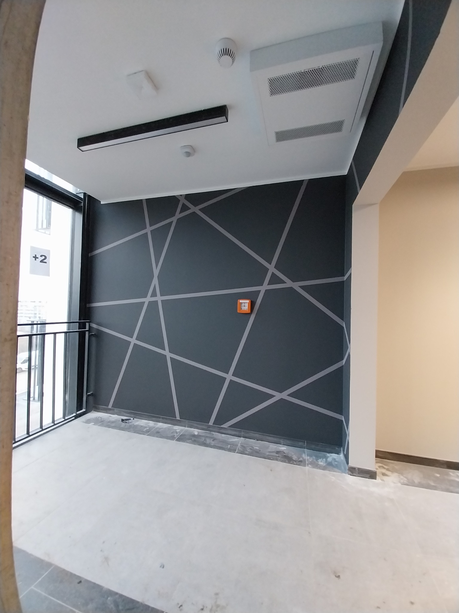 Wnętrze w stanie deweloperskim z szarą ścianą zdobioną geometrycznym wzorem z jasnych linii, widoczny fragment balkonu, sufit z oświetleniem liniowym i kratką wentylacyjną.