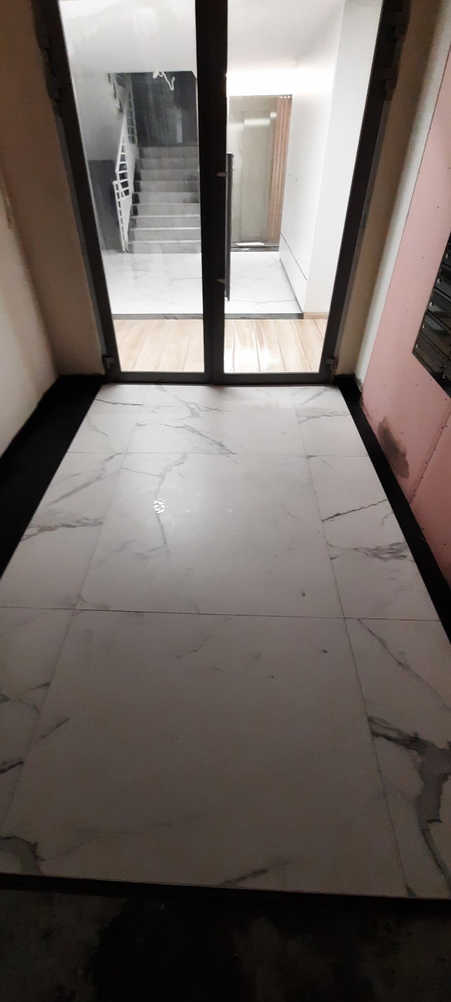 Świeżo położone, białe płytki podłogowe z marmurkowym wzorem, widoczne przez przeszklone drzwi wejściowe, z widokiem na schody w oddali.