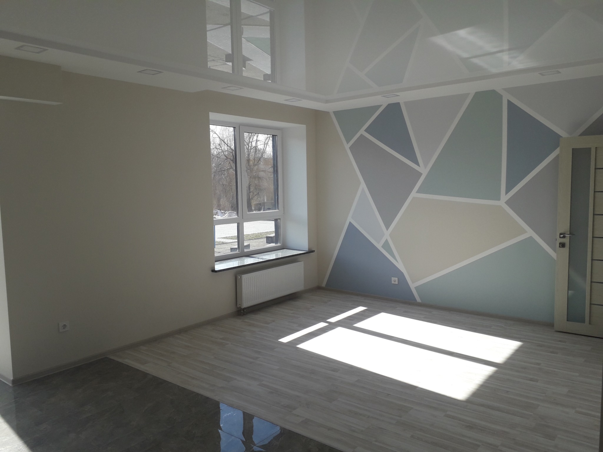 Jasne, przestronne wnętrze pokoju po odświeżeniu, z geometrycznym wzorem na ścianie w odcieniach szarości i błękitu, panelami podłogowymi imitującymi drewno oraz lśniącym, podwieszanym sufitem...