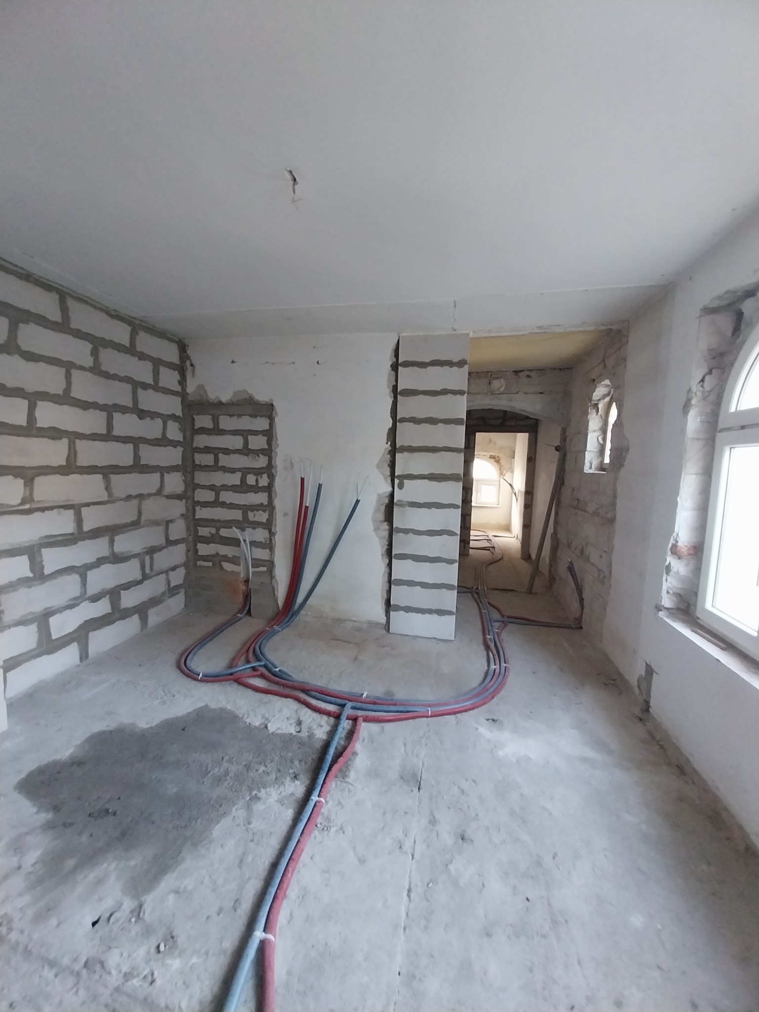 Wnętrze w trakcie prac: surowe ściany z bloków, częściowo otynkowane, z widocznymi instalacjami wodnymi (czerwone i niebieskie rury), surowy beton na podłodze z plamami, przygotowywane do dalszych...