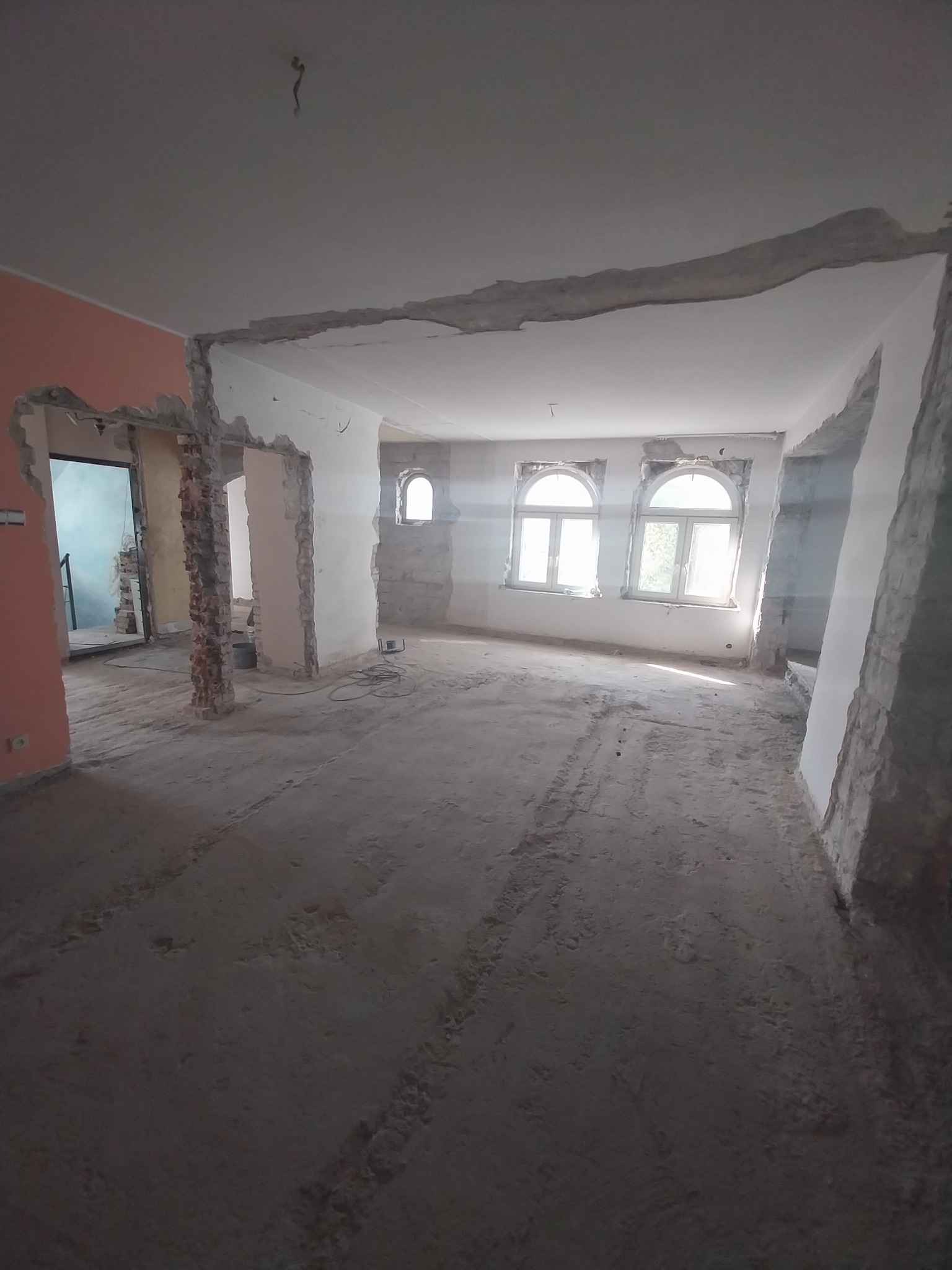 Przestronne wnętrze w trakcie remontu: usunięte ściany działowe, odsłonięta cegła przy otworach drzwiowych, resztki tynku na ścianach i suficie, przygotowanie do dalszych prac wykończeniowych.