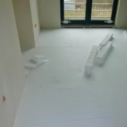 Białe płyty styropianowe rozłożone na podłodze w surowym stanie, przygotowane pod wylewkę anhydrytową, widoczne przycięte fragmenty i okno w tle.