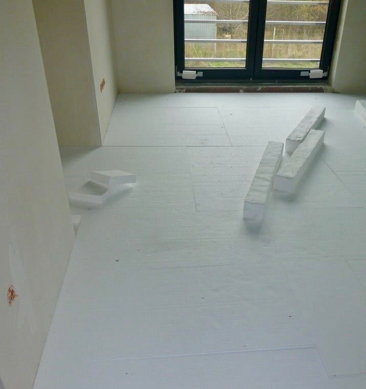 Białe płyty styropianowe rozłożone na podłodze w surowym stanie, przygotowane pod wylewkę anhydrytową, widoczne przycięte fragmenty i okno w tle.