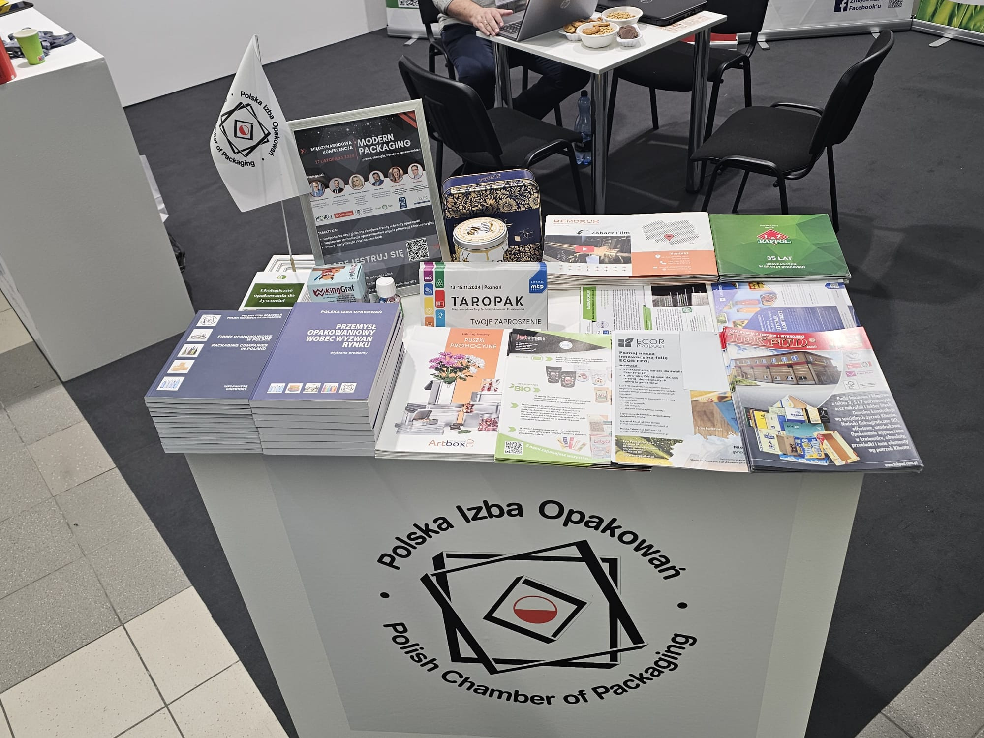 Stoisko targowe Polskiej Izby Opakowań z ulotkami, zaproszeniami i materiałami promocyjnymi, widoczny laptop i przekąski na stole w tle.